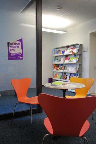roter, oranger und gelber Stuhl, um einen kleinen runden Marmortisch gruppiert, im Hintergrund ein Regal mit Informationsbroschüren